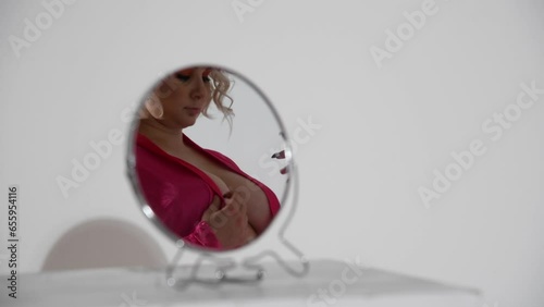 Small round mirror stands on white table in white room. Sexy caucasian plus size woman wearing pink robe on naked body reflects on mirror surface. Real time video. Abstract sensual background.
