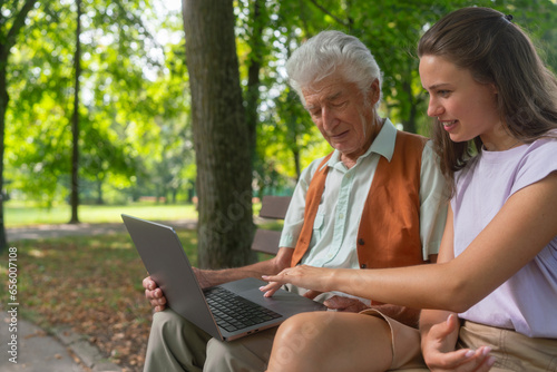 The grandfather and granddaughter are looking at photos on notebook, sitting on a bench in a city park. photo