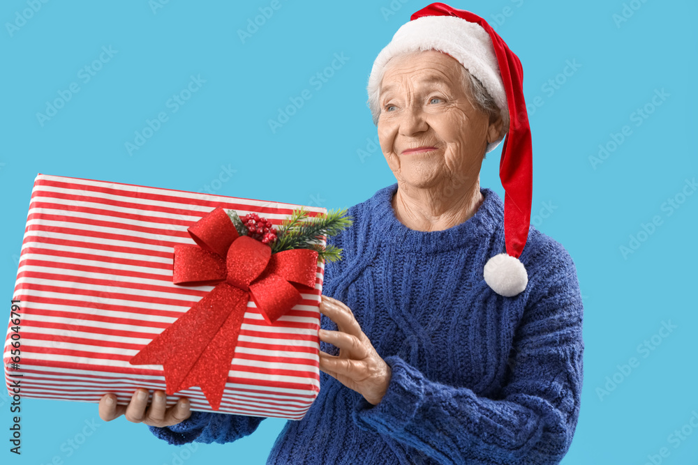 Obraz premium Senior woman in Santa hat with Christmas gift on blue background