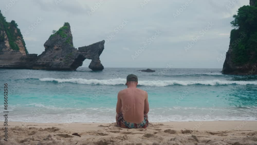 Anonymous man sitting on ocean shore watching tidal waves washing sand beach against fabulous landscape with dark rocks. Background with seaside and tourist. Turquoise waves in summer. Travel tropics. Anonymous man sitting on ocean shore watching tidal waves washing sand beach against fabulous landscape with dark rocks. Background with seaside and tourist. Turquoise waves in summer. Travel tropics.