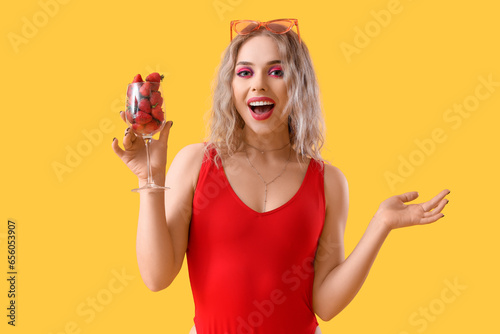 Young woman in swimsuit with glass of strawberries on yellow background