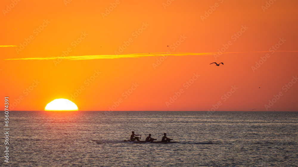 Boat with three people, a seagull flying around and with the sun almost gone in the horizon at sunset