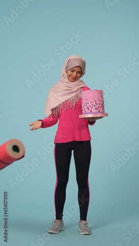 Full-sized isolated photo capturing a young woman in a sportswear and a hijab, sheila choosing between a cake and an exercising mat, throwing a mat away. photo