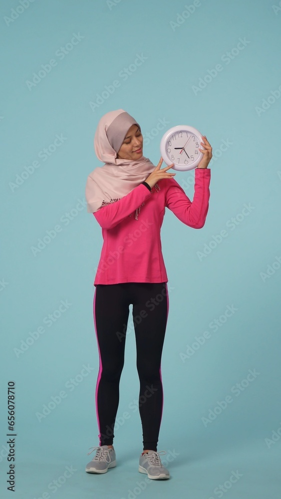 Full-sized isolated photo capturing an attractive young woman wearing a sportswear and a hijab, sheila. She is holding a clock and smiling. Full-sized isolated photo capturing an attractive young woman wearing a sportswear and a hijab, sheila. She is holding a clock and smiling.
