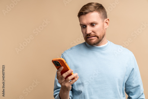 Scared frustrated man reding message in smartphone looking at screen isolated on beige background. Bearded middle aged guy having bad news, problems, troubles. Male feeling confusion, desperation. © DimaBerlin