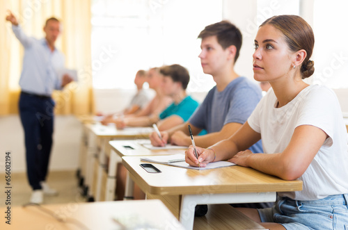 Portrait of concentrated attractive teenage girl studying at university, listening attentively to lecturer and writing in workbook