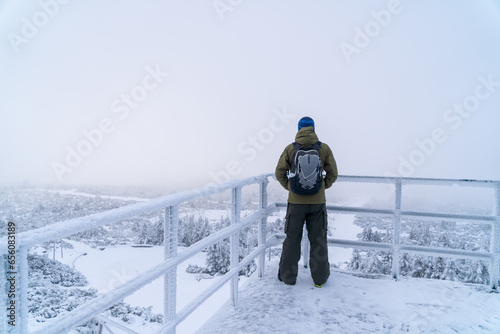 Frost on the railing of the metal fence. Man in winter clothes with backpack stand on a edge of view point and look down on the background of a snow forest with rocks.