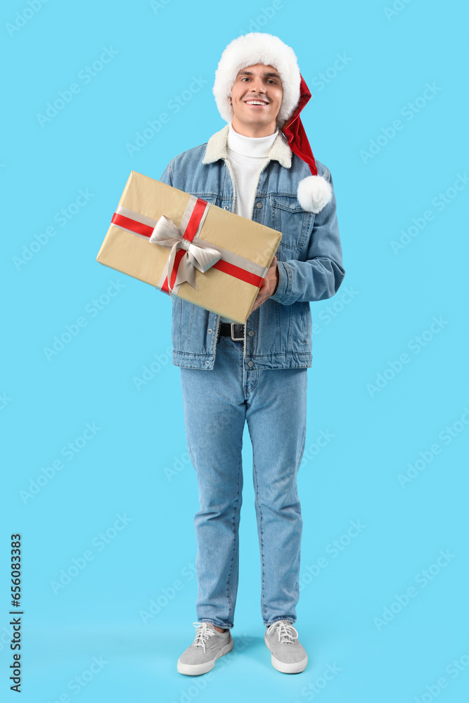 Happy young man in Santa hat with gift box on blue background Happy young man in Santa hat with gift box on blue background