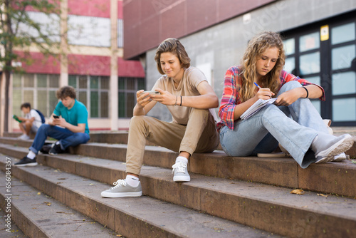 Teenager girl sitting on stairs beside school building and doing homework. Her classmate sitting next to her and using smartphone.