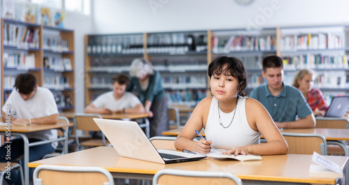 Fifteen-year-old Asian schoolgirl is studying in the school library on a laptop while preparing for an exam, making important notes in a copybook