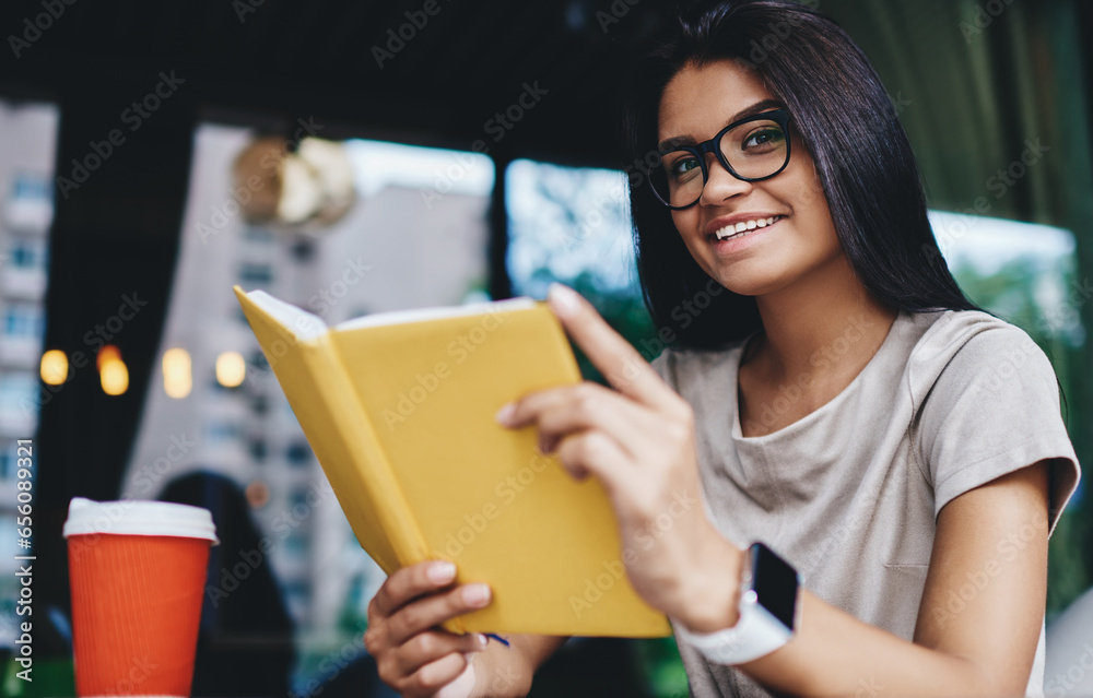 smiling hipster girl in eyewear enjoying new best seller looking at camera in cafe smiling hipster girl in eyewear enjoying new best seller looking at camera in cafe
