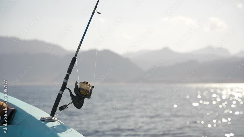 Close up of fishing rod with the reel in holder on a motor boat in the sea at sunset. Mountains on the coast in the background Close up of fishing rod with the reel in holder on a motor boat in the sea at sunset. Mountains on the coast in the background