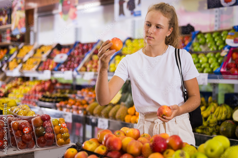 Portrait of teenage blonde girl customer buying sweet golden apples at grocery shop