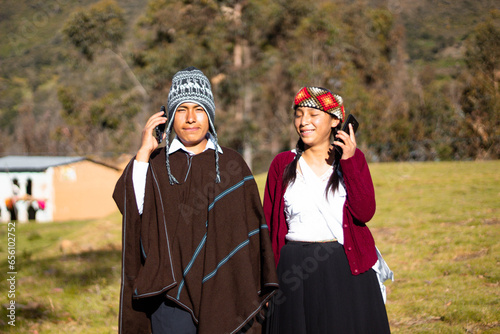Hombre nativo quechua usando un chullo, poncho y teléfono a lado de una mujer atractiva sosteniendo celular en la mano, aire libre, comunicación