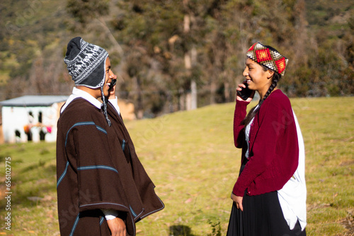 Hombre nativo quechua usando un chullo, poncho y teléfono a lado de una mujer atractiva sosteniendo celular en la mano, aire libre, comunicación photo