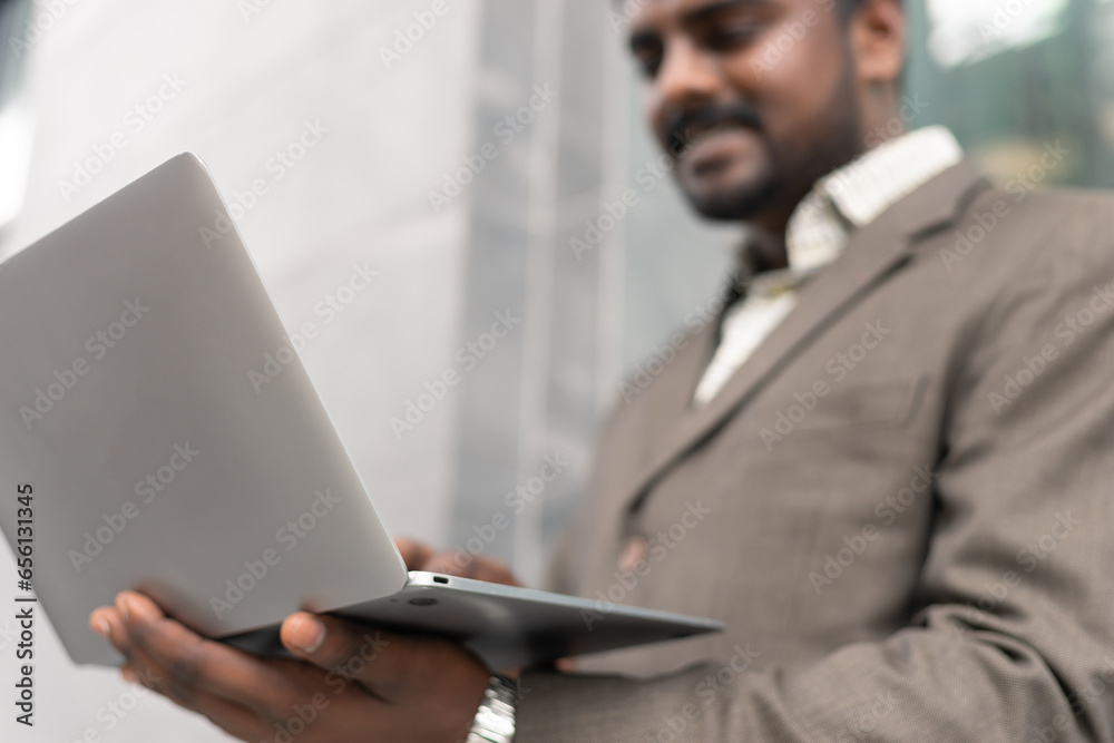 Young Indian businessman smiling and using the laptop nearby his office workspace against contemporary corporate buildings in the city. Young Indian businessman smiling and using the laptop nearby his office workspace against contemporary corporate buildings in the city.