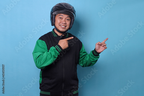 Smiling Asian online taxi driver with safety helmet and uniform jacket pointing aside to empty space for text or advertisement