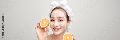 Young woman applying clay mask on her mask with sliced fruit in hand over color banner