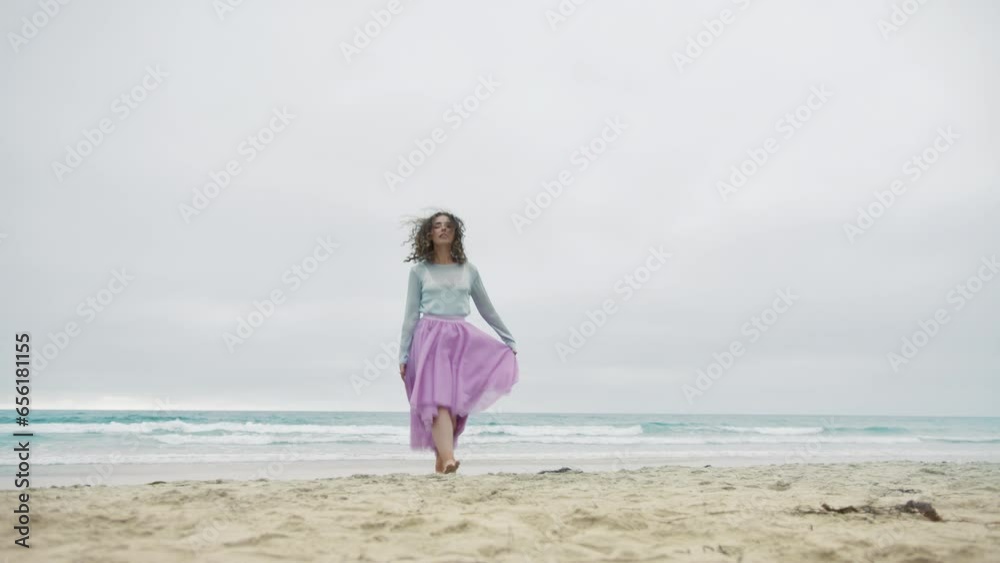 Happy cheerful girl in pink skirt jumping on ocean beach on summer cloudy day. Attractive curly woman running by sand waving hands in dance. Smiling cheerful lady have fun, enjoying beautiful nature Happy cheerful girl in pink skirt jumping on ocean beach on summer cloudy day. Attractive curly woman running by sand waving hands in dance. Smiling cheerful lady have fun, enjoying beautiful nature