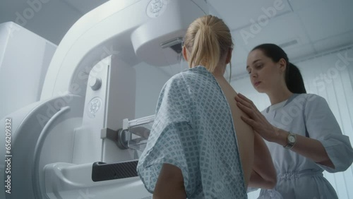 Hospital radiology room. Caucasian woman stands during mammography screening examination in clinic. Female doctor adjusts modern mammogram machine for patient, uses computer. Breast cancer prevention.