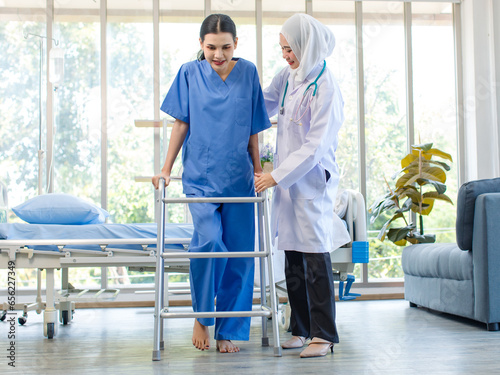 Asian professional successful experienced muslim female doctor in white lab coat with hijab stethoscope helping supporting assisting injury patient walking using four legs walker in hospital ward