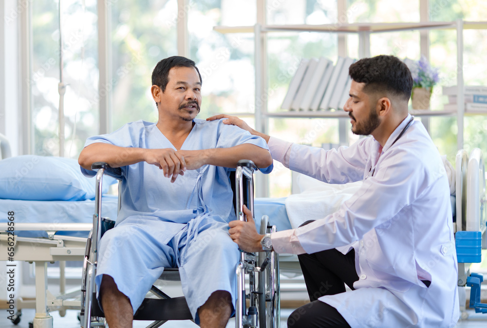 Asian Indian professional young handsome bearded male doctor in white lab coat uniform stethoscope visiting helping supporting assisting injury senior male patient on wheelchair in hospital ward Asian Indian professional young handsome bearded male doctor in white lab coat uniform stethoscope visiting helping supporting assisting injury senior male patient on wheelchair in hospital ward