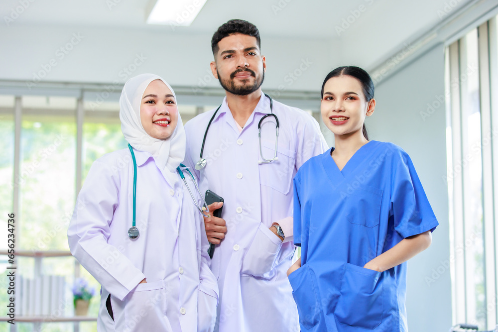Asian Indian professional experienced bearded male doctor in white lab coat with stethoscope with female muslim colleague in hijab and surgeon in blue uniform standing smiling together in hospital Asian Indian professional experienced bearded male doctor in white lab coat with stethoscope with female muslim colleague in hijab and surgeon in blue uniform standing smiling together in hospital