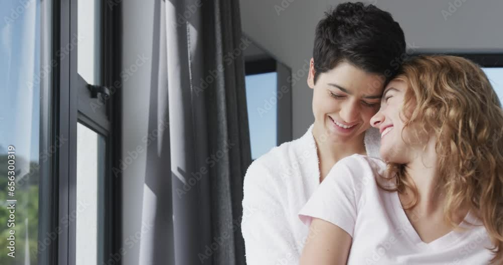 Happy caucasian lesbian couple standing by window, embracing and smiling in sunny house Happy caucasian lesbian couple standing by window, embracing and smiling in sunny house