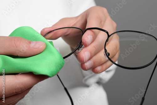 Woman wiping her glasses with microfiber cloth on grey background, closeup
