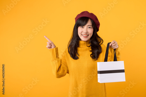 Young Asian woman in her 30s, with an elegant flair, holding a shopping paper bag, pointing her finger, donning a yellow sweater and red beret against a yellow background. photo