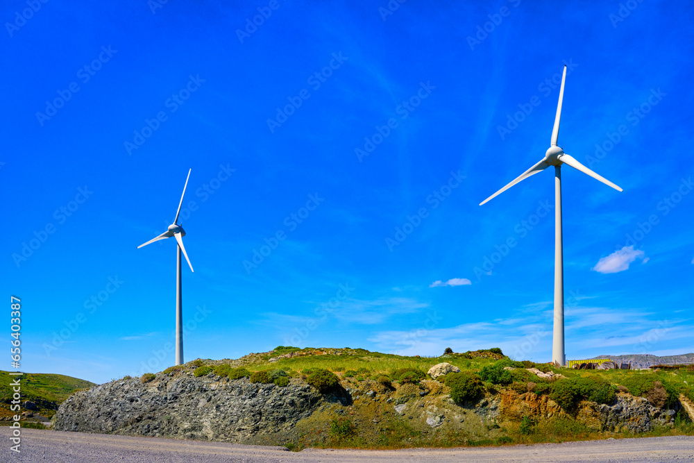 Two wind turbines by road on top of green hill on sunny day, Greece
