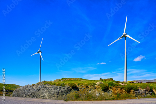 Obraz na płótnie Two wind turbines by road on top of green hill on sunny day, Greece w salonie