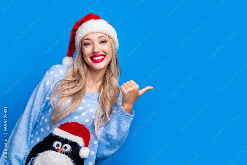 Obraz premium Photo portrait of attractive blonde young woman smile point empty space wear penguin sweater x-mas hat isolated on blue color background