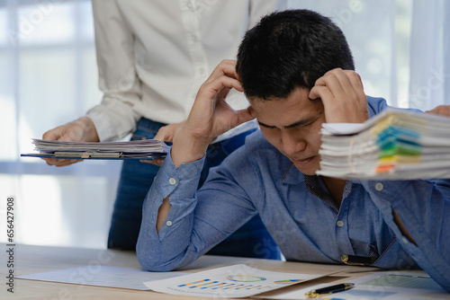 Stressed businessman sitting at workplace and touching his headYoung asian exhausted businesssman with messy desk and stack of papers, working busy, overwork. photo