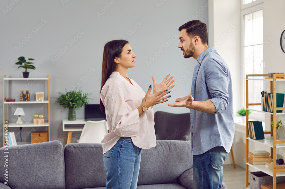 Emotional stressed young couple having argument at home. Portrait of angry irritated man and woman talking and looking at each other with annoyed. Relationship problems, family conflicts - obrazy, fototapety, plakaty 