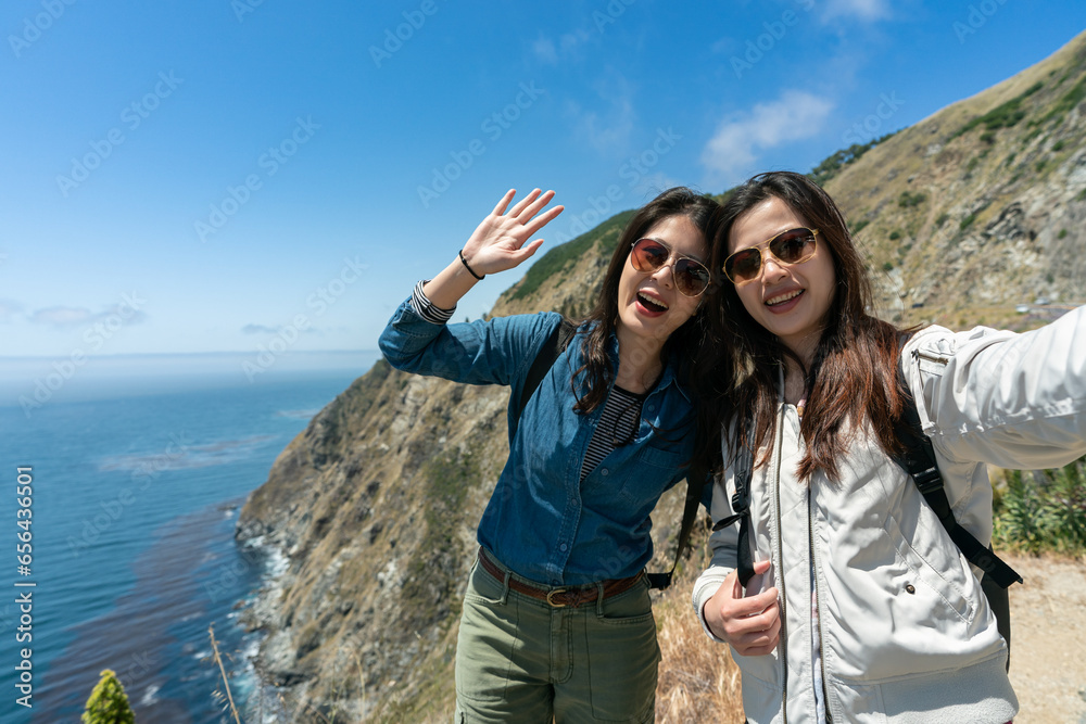 two smiling asian Japanese female tourist taking selfie picture on backdrop of ocean and rugged hill on sunny day. one is waving palm to camera