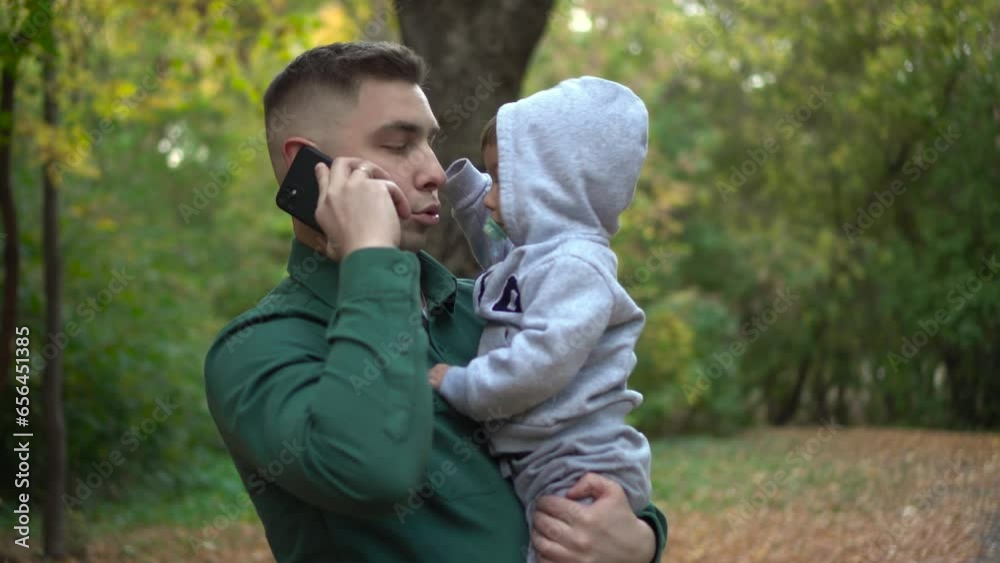 A young father holds his son in his arms and talks on the phone. A man with a child in the park with a smartphone. A young father holds his son in his arms and talks on the phone. A man with a child in the park with a smartphone.