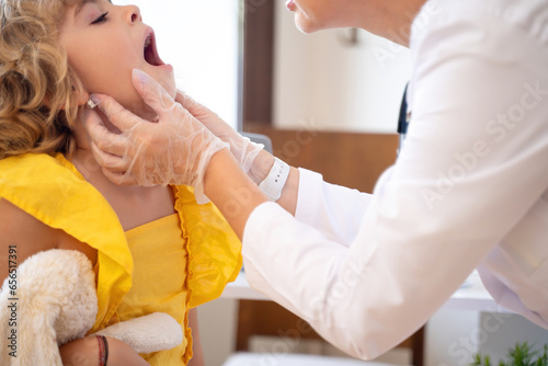 Female doctor checking child's exam kid throat during medical checkup in medical exam room at the clinic. Friendly pediatrician using stethoscope to examine breathing and heartbeat of young patient photo