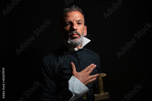 Man dressed in black and white collar with serious gesture that looks like the man from El Greco's Knight of the Hand on the Chest on a black background