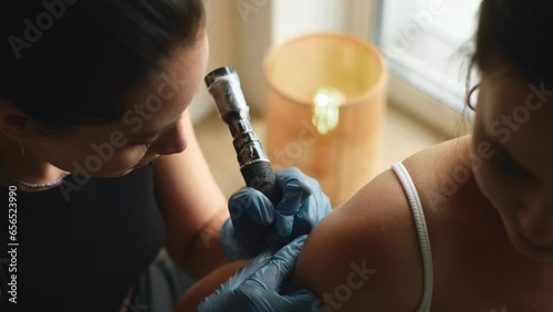 Young woman in tattoo salon during procedure. Professional female tattooist makes tattooing on customers shoulder in tattoo studio. Cinematic shot