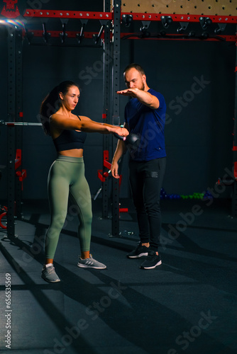 beautiful young fitness woman doing exercises with dumbbells in hands with trainer in gym preparing for bodybuilding competition