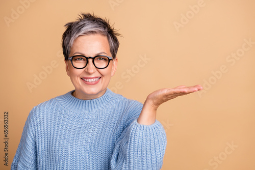 Photo portrait of lovely retired female palm hold empty space dressed stylish blue knitted outfit isolated on beige color background