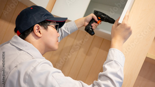 Asian male furniture assembler using electric drill screwdriver on stainless steel cabinet hinge. Interior construction worker man using screwdriving tools for furniture installation in the new house.