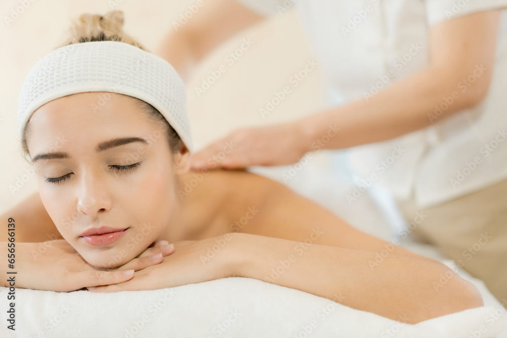 Beautiful young woman enjoying professional wellness massage in spa salon, woman masseur massaging back and shoulder blades of young woman lying on massage table on white background With Closed Eyes.