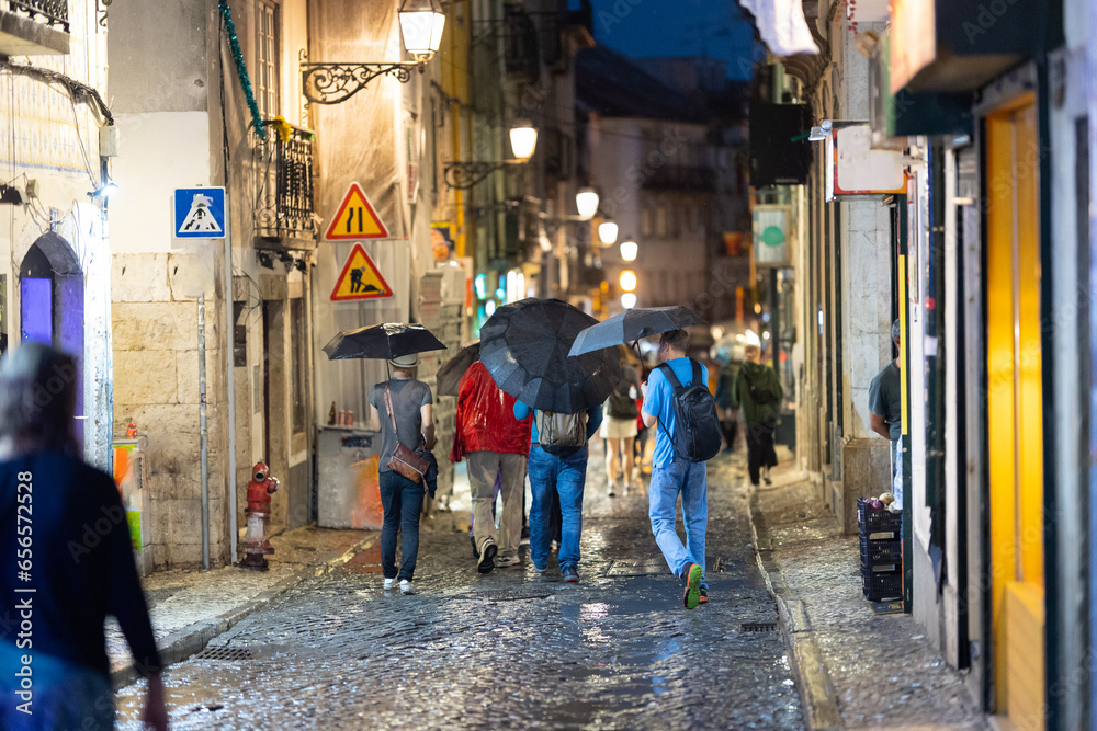 People walk along a narrow European street with umbrellas in the rain - colored lights of the night city