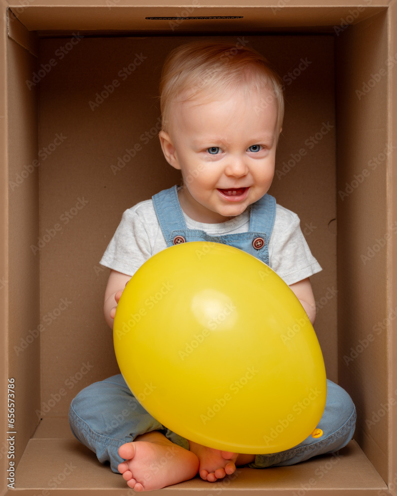Cute baby boy playing with yellow ballon sitting in cardboard box. Child with toy. Cute baby boy playing with yellow ballon sitting in cardboard box. Child with toy.