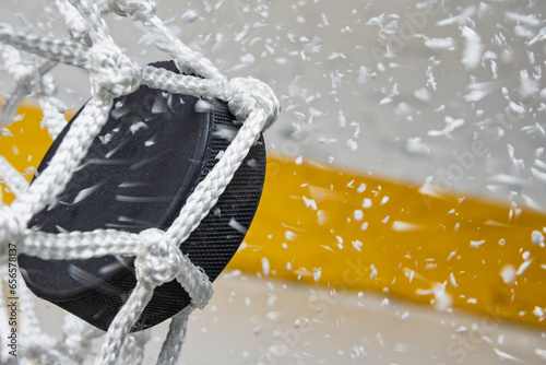 A close-up view of an Ice Hockey puck hitting the back of the goal net as shavings fly by, viewed from the side. Scoring a goal in ice hockey.