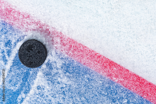 Looking down on a black ice hockey puck plowing through snow and stopping on the edge of the Goal Line of the goal crease.  photo