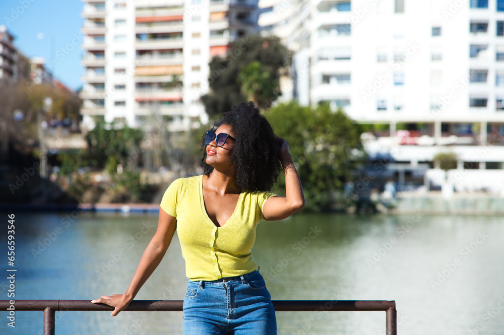Young, pretty black woman with afro hair and sunglasses wearing jeans and yellow shirt leans on the railing. In the background the river guadalquivir and the neighbourhood in seville.