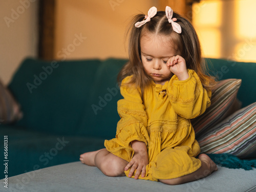Photos Upset sad sleepy little toddler girl sitting alone on the sofa and rubbing her eyes
